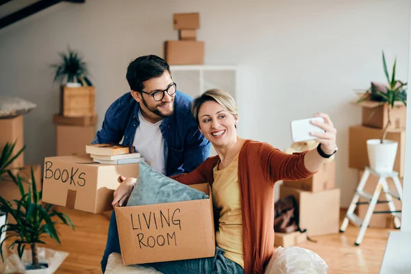 Happy couple using smart phone and taking selfie while moving into new apartment.