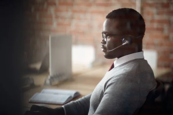 Side view of black businessman working on desktop PC and wearing headset in the office. The view is through the glass. Copy space.