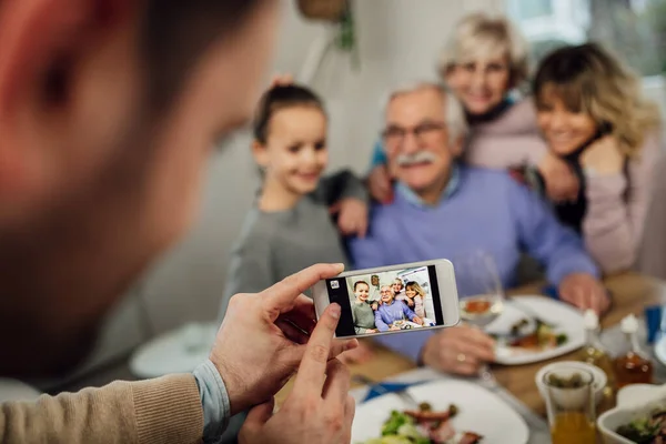 Close-up of man using smart phone while photographing his happy extended family in dining room.