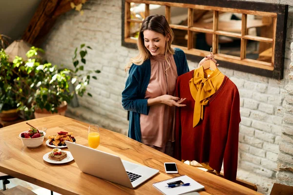 Happy woman showing her clothing selection to someone during video call over laptop at home.