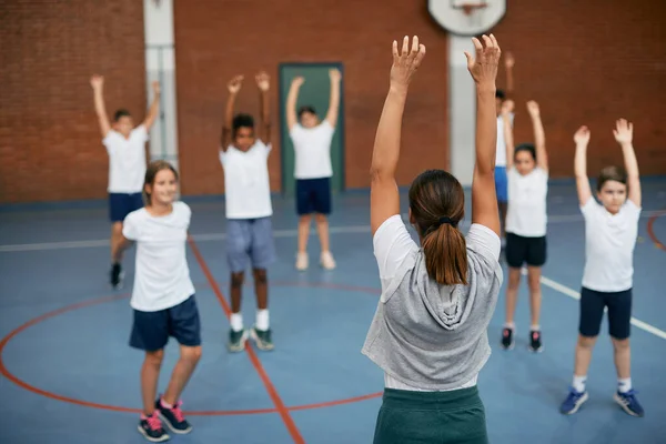 Back view of PE teacher and group of elementary students exercising during a class at school gym. 