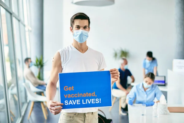 Young man with face mask holding placard and inviting people to get vaccinated against COVID-19 and save lives.
