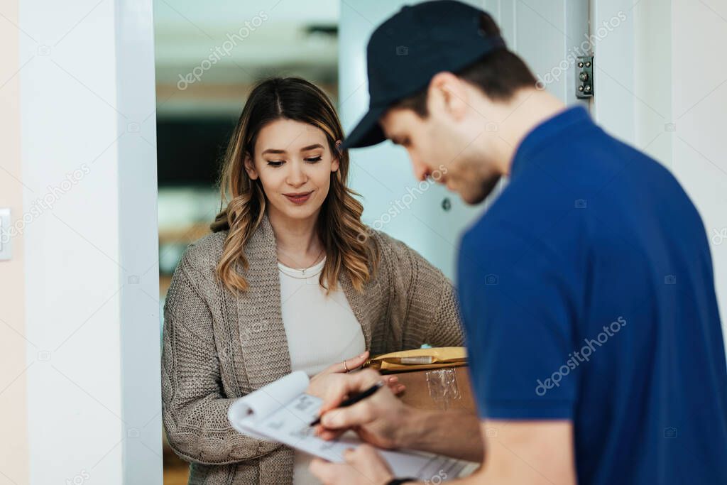 Smiling woman receiving package and waiting for a delivery man to fill paperwork while standing on a doorway. 