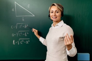 Smiling teacher giving math lesson online while explaining it on chalkboard in the classroom. 