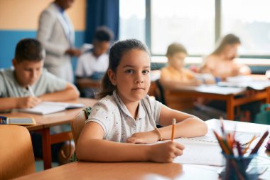 Cute schoolgirl writing in notebook during a class in the classroom and looking at camera. 