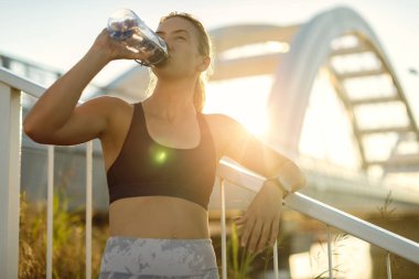 Young female athlete drinking water from a bottle while taking a break from exercising outdoors. 