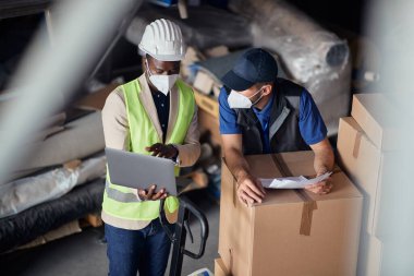 Black foreman and warehouse worker talking while using laptop and wearing protective face masks storage compartment. 