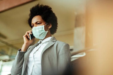 African American businesswoman talking on smart phone while walking through airport corridor with protective mask on her face. Copy space. 
