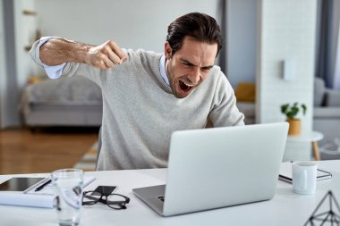 Angry businessman shouting from frustration and about to punch his laptop after reading bad news while working at home. 