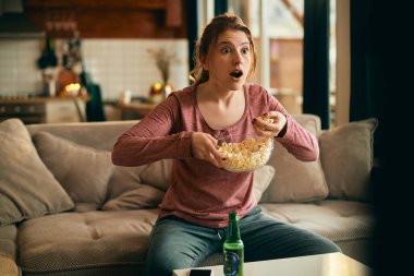 Young woman watching something shocking on TV while eating popcorn in the living room. 