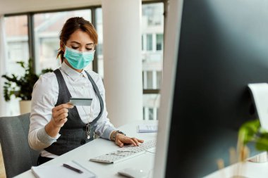 Female entrepreneur with face mask using computer and credit card while online shopping in the office. 