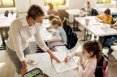 Elementary school teacher helping her student in learning in the classroom. They are wearing protective face masks due to coronavirus pandemic. 