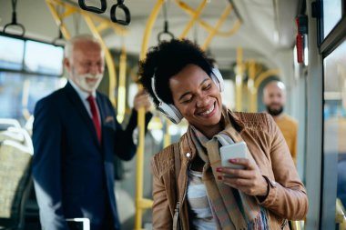 Happy African American using mobile phone and listening music over headphones while commuting by public transport. 