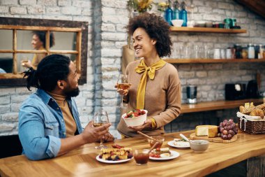 Happy African American woman offering her husband strawberries while drinking wine and talking at home. 