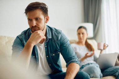 Displeased man thinking of something while having problems with his wife who is using laptop in the background.