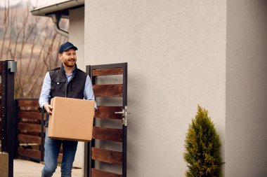 Smiling male courier carrying cardboard box while making home delivery to his customer. 