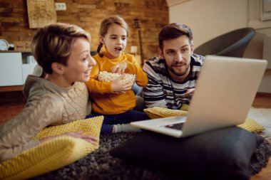 Happy small girl eating popcorn while using laptop with her parents at home. 