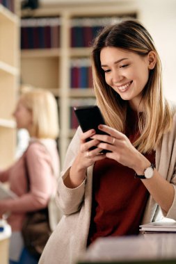 Happy female student using smart phone while being at university library. 