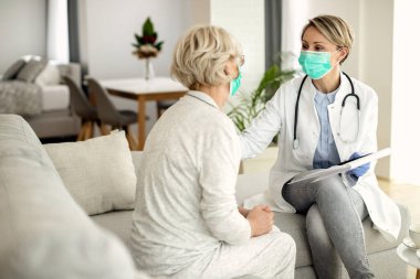 Female doctor and senior woman talking during home visit and wearing protective face masks due to coronavirus pandemic. 