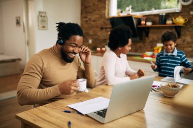 Happy African American man using laptop and making video call while drinking tea and working at home. His wife and son are in the background. 