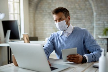 Businessman wearing protective face mask while working on a computer and using digital tablet in the office. 