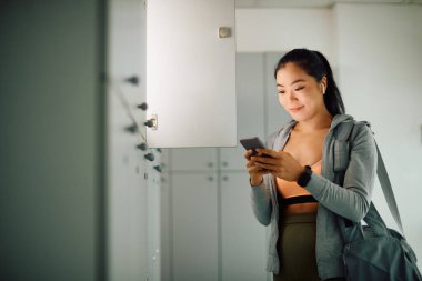 Genç Asyalı bayan sporcu spor salonundaki soyunma odasında cep telefonu ve kısa mesaj kullanıyor..
