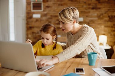Mid adult mother using laptop and assisting her daughter with e-learning at home. 