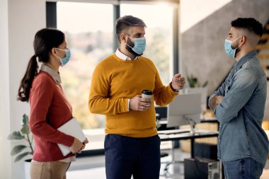 Group of entrepreneurs wearing protective face masks while communicating on a break in the office. Focus is on man in yellow sweater. 