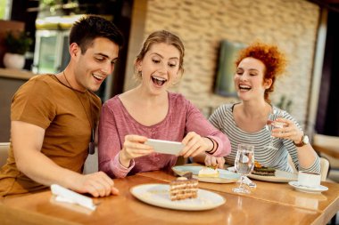 Young happy friends eating dessert and having fun while taking picture of it in a cafe, 