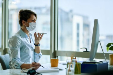 Businesswoman with face mask working on a computer while recording voice message on smart phone in the office. 