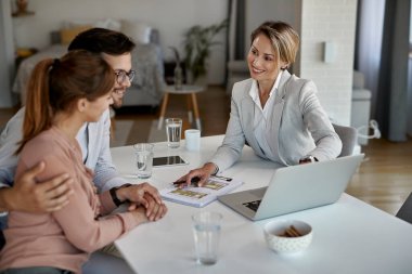 Happy real estate agent and young couple talking while using laptop during the meeting. 