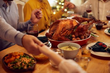 Close-up of family holding hands and praying while having Thanksgiving lunch at home. Focus is on roast turkey.