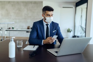 Male entrepreneur working on computer while using mobile phone and wearing face mask due to COVID-19 epidemic. 