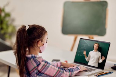 Little girl with face mask using computer and listening to her teacher who is holding online class due to coronavirus pandemic. 