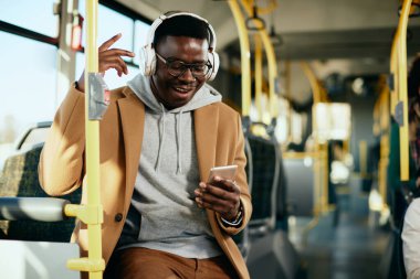 Happy black man having fun while listening music over headphones and using mobile phone in a bus. 