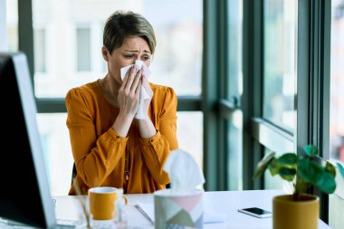 Businesswoman feeling sick and blowing her nose while working in the office.