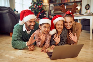 Happy parents and their daughters using laptop and greeting someone during video call on Christmas.