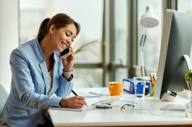 Young happy female entrepreneur taking notes and talking on mobile phone while working the office. 