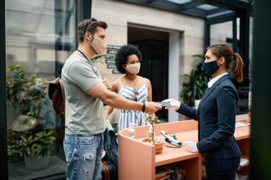 Happy man and his wife checking in at a hotel and giving passport to receptionist while traveling during COVID-19 pandemic.