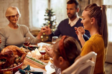 Mid adult woman and her family saying grace and holding hands during Christmas meal in dining room. 