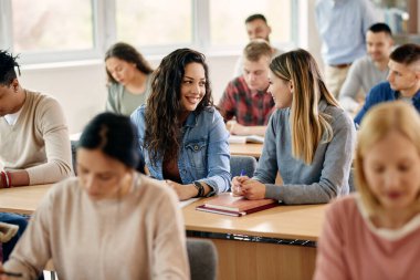 Happy college student and her female friend communicating while attending a class at lecture hall. 