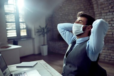 Young businessman wearing protective face mask and relaxing with hands behind head in the office.