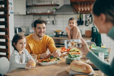 Mother using smart phone and photographing her husband and daughters while they are setting dining table for lunch in the kitchen. Focus is on man. 