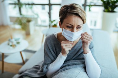 Worried woman wearing face mask and thinking while sitting at home during coronavirus lockdown. 