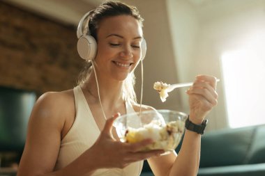Happy athletic woman eating salad while listening music over headphones and relaxing at home. 