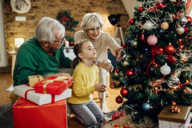 Happy mature couple with granddaughter decorating Christmas tree in the living room. 