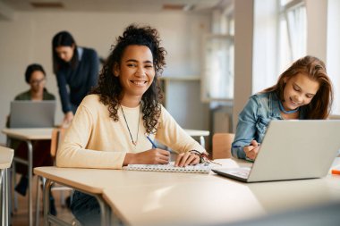 Happy African American high school student writing notes while e-learning on laptop with her classmate in the classroom. 