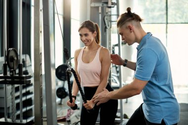 Happy athletic woman doing rope push-downs while having sports training with fitness instructor in a gym.