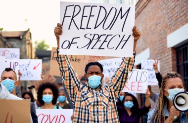 African American man wearing protective face mask and holding a placard with 'freedom of speech' inscription while protesting on city streets. 