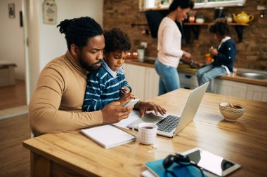 Black working father using laptop and mobile phone while son is sitting on his lap. Mother and daughter are in the background. 